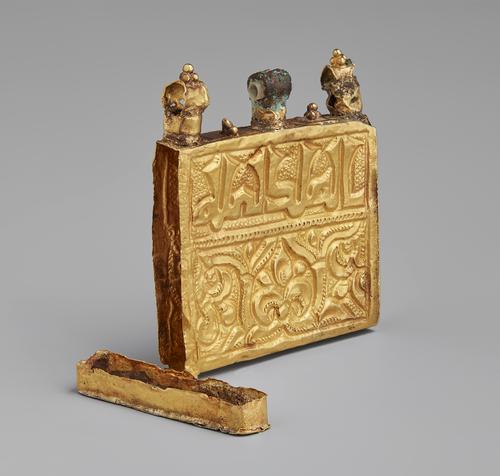 Side view of the gold amulet qur’an case. Square gold thin box with three loops on the top where it would be hung as a pendant. Inscription and engraving on the front panel and the side cover is open to see the hollow interior.