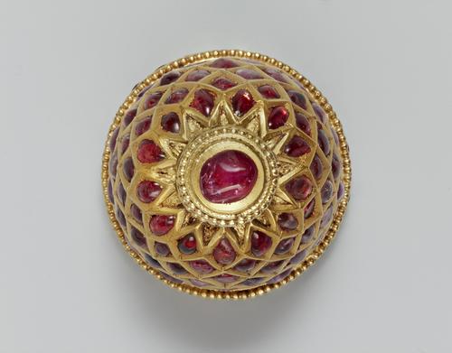 Top view of the small jewelled container encrusted with red gemstones. Its top, which is surmounted with a red cabochon stone and surrounded by small gold granules. 