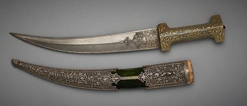 Dagger with jade hilt with golden arabesques and a steel blade with an inscription closest to the hilt. Scabbard is seen beside it, is decorated with metal floral patterns at the tip and top, with green velvet like fabric in the middle section. 