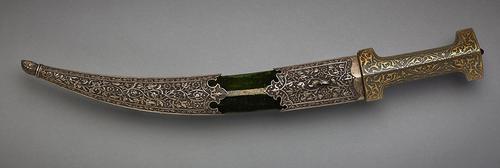 View of dagger with jade hilt with golden arabesques inside the scabbard which is decorated with metal floral patterns at the tip and top, with green velvet like fabric in the middle section.