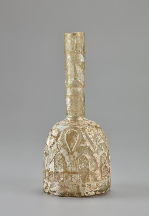 Side of a Bottle of cylindrical form with rounded shoulder and tall tubular neck on short foot, the body has a faceted pattern, the shoulder cut with inverted tear-drop motifs, the neck with rectangular facets and cut with a zigzag design, white iridescence. 