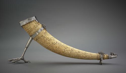 Elephant tusk, conical-shaped with surface decoration of horizontal bands along which processions of animals and birds run after each other. Silver band at the mouth of the horn and a silver claw resembling a bird’s foot underneath which acts as a stand.