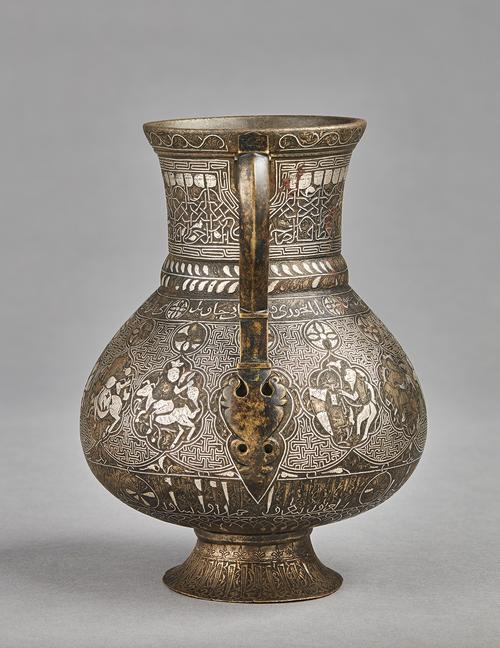 A pear-shaped brass jug with handle on a gently splayed foot, with silver inlaid decoration comprising of interlinked medallions containing representations of the zodiac signs, on a T-fret pattern background, between two inscriptive bands; the neck with a frieze of human-headed Kufic inscription. 