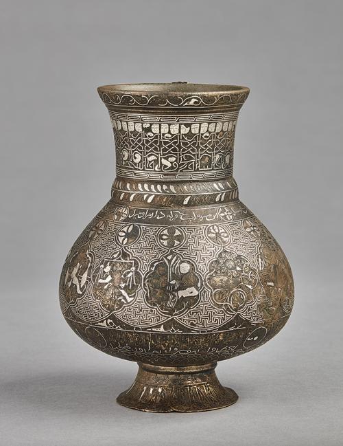 A pear-shaped brass jug on a gently splayed foot, with silver inlaid decoration comprising of interlinked medallions containing representations of the zodiac signs, on a T-fret pattern background, between two inscriptive bands; the neck with a frieze of human-headed Kufic inscription. 