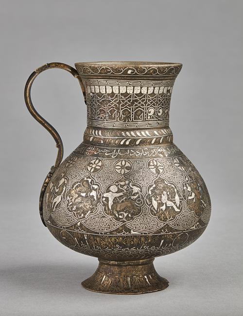 A pear-shaped brass jug with handle on a gently splayed foot, with silver inlaid decoration comprising of interlinked medallions containing representations of the zodiac signs, on a T-fret pattern background, between two inscriptive bands; the neck with a frieze of human-headed Kufic inscription. 