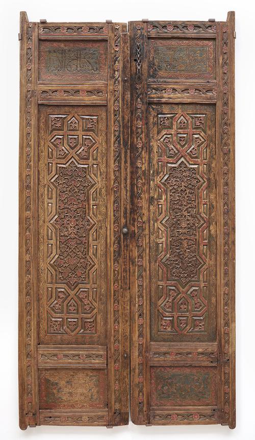 A double wooden door. Each rectangular door is decorated with inset panels deeply carved and painted in polychrome, high reliefs of floral motifs, and shallow-carved inscriptions. 