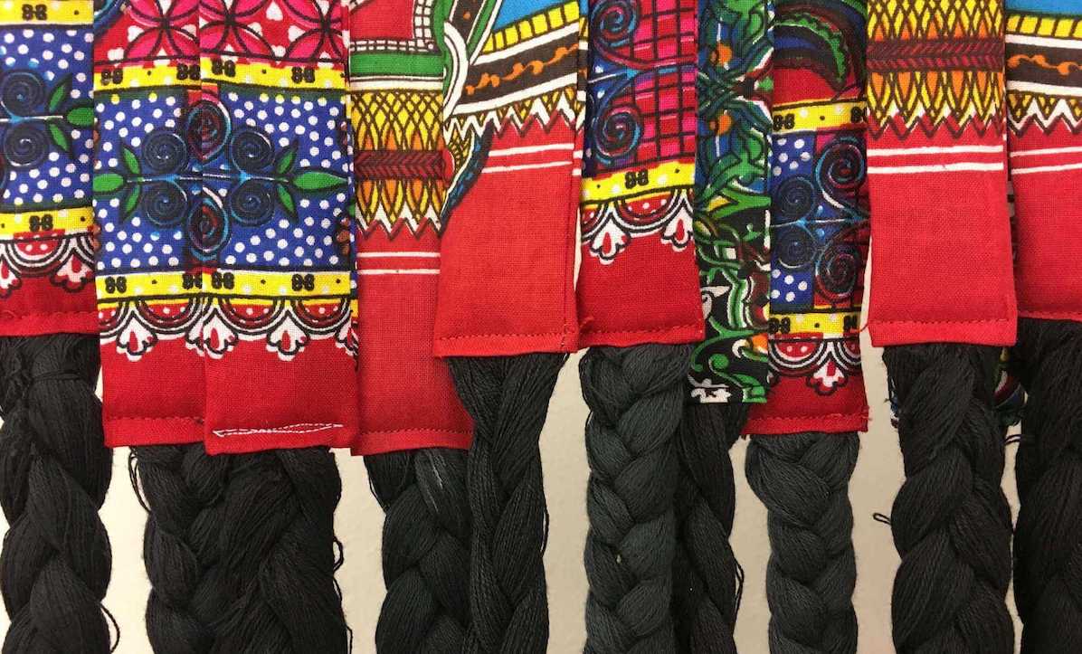 Braids of black yarn are suspended below strips of textile with a red pattern.