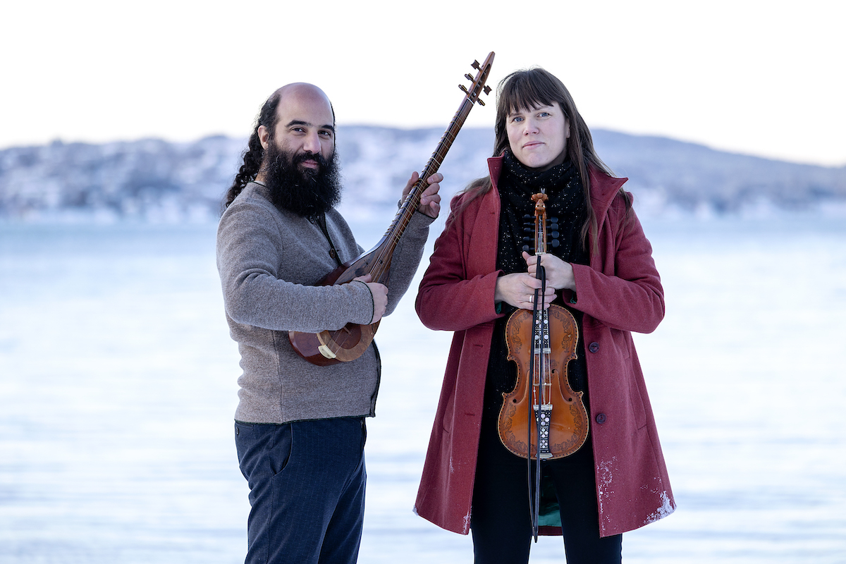 Kiya Tabassian stands in an icy terrain next to Benedicte Maurseth holding his sitar while she stands holding her fiddle. 