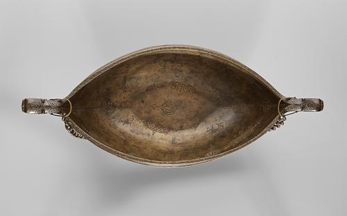 Top view of a narrow brass bowl, showing the interior which is engraved with geometric designs. At each pointed end of the bowl is a small dragon head, from which a chain hangs.